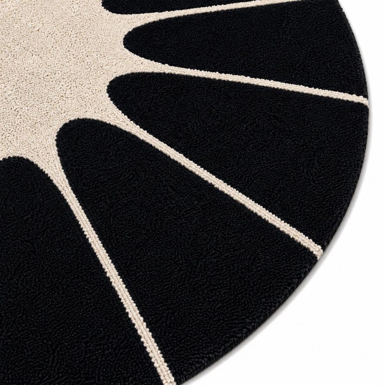 May include: A circular rug featuring a high-contrast design with a central cream-colored sunburst pattern radiating outward into deep black segments. The textured surface displays a dense, tufted pile construction with clean, defined lines.