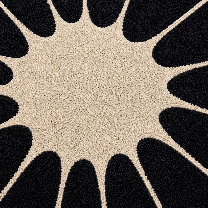 May include: A circular rug featuring a textured, tufted surface with a high-contrast design. The center is a solid cream-colored circle, surrounded by radiating, petal-like shapes in black that extend outward to the edge of the rug.