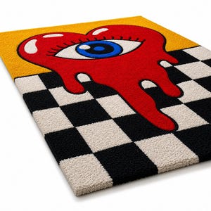 May include: A rectangular tufted rug featuring a large red heart with a central blue eye and long eyelashes. The heart appears to be melting downward over a black and white checkered pattern, set against a solid yellow background at the top.