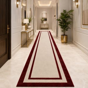 May include: A long rectangular runner rug featuring a solid cream-colored center panel framed by two distinct, bold dark red borders. The rug is positioned centrally along a polished light-colored floor in a bright, symmetrical hallway interior.