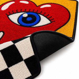 May include: A rectangular tufted rug featuring a large red heart with a blue eye in the center against a yellow background. The bottom section displays a black and white checkered pattern. The rug has a black bound edge and a black textured backing.