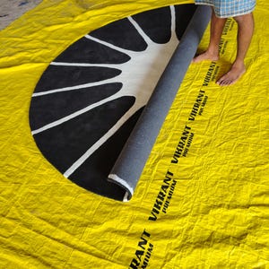 May include: A semi-circular rug featuring a black background with a white sunburst pattern. The rug is partially rolled, revealing a dark gray backing. The rug is placed on a yellow tarp printed with the text VIKRANT PREMIUM.