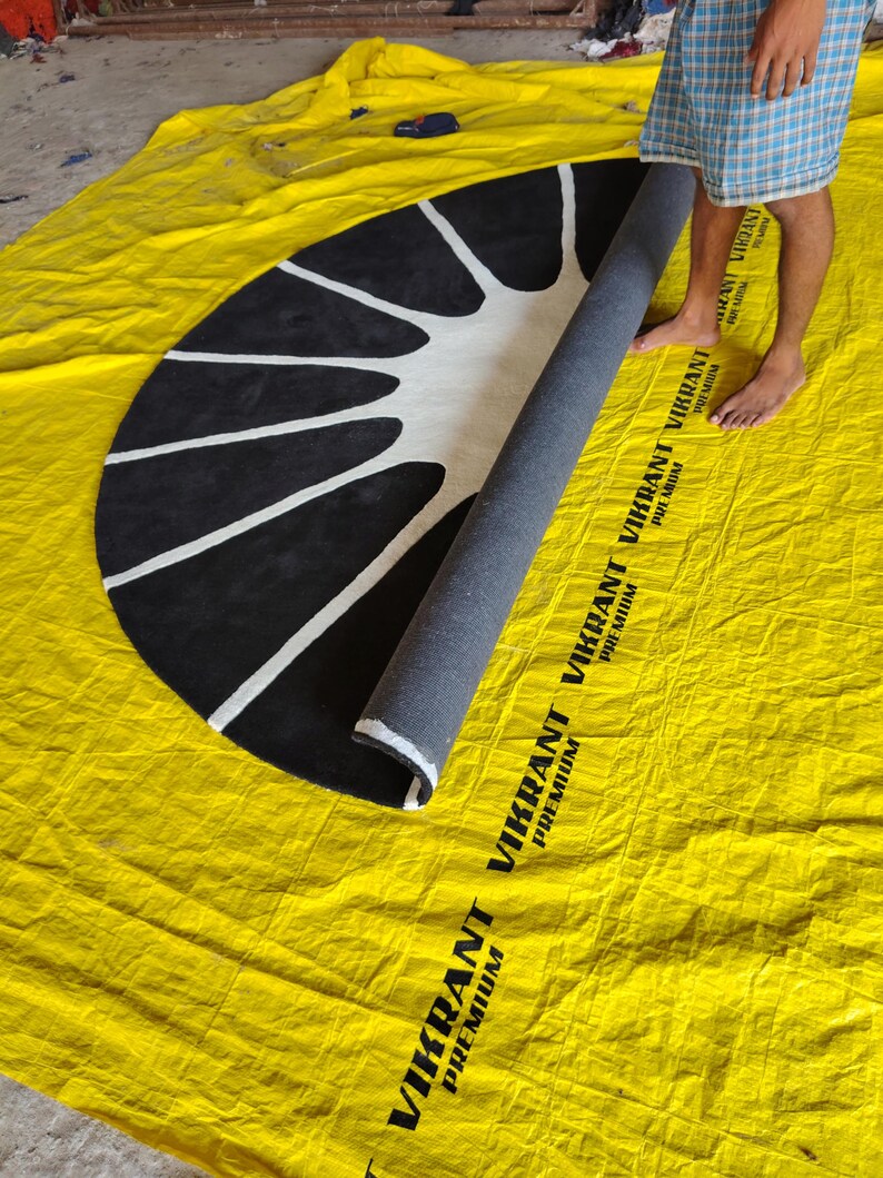 May include: A semi-circular rug featuring a black background with a white sunburst pattern. The rug is partially rolled, revealing a dark gray backing. The rug is placed on a yellow tarp printed with the text VIKRANT PREMIUM.