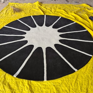 May include: A circular rug featuring a high-contrast design with a solid black background and a central white star-like shape. The white pattern extends from the center toward the outer edge in twelve distinct, elongated, tapered rays.