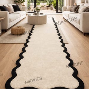 Könnte beinhalten: Ein cremefarbener Teppich mit schwarzem Rand und gewelltem Rand. Der Teppich liegt auf einem Holzboden in einem Wohnzimmer, zusammen mit weißen Sofas und einem runden Couchtisch. Das Design des Teppichs verleiht dem Raum eine moderne Note.
