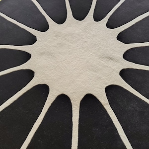 May include: A circular rug featuring a high-contrast design with a central off-white starburst shape that extends outward into long, thin, curved points, set against a solid black background that fills the remaining space of the circle.