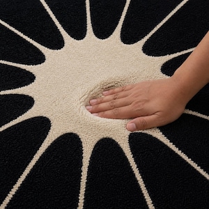 May include: A circular rug featuring a high-contrast design with a central beige sunburst pattern radiating outward into a deep black background. The textured, tufted surface shows a visible indentation where pressure is applied to the material.