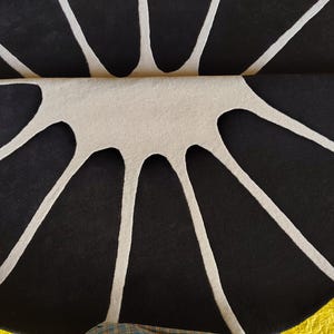 May include: A circular rug featuring a high-contrast pattern of black segments separated by white lines that radiate from the center. The rug is folded in half, revealing a textured surface with a distinct geometric design.
