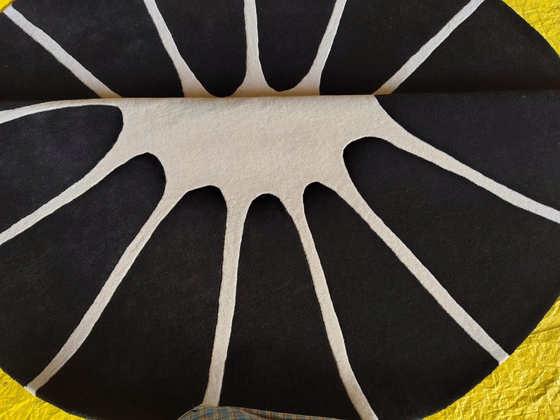 May include: A circular rug featuring a high-contrast pattern of black segments separated by white lines that radiate from the center. The rug is folded in half, revealing a textured surface with a distinct geometric design.