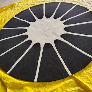 May include: A circular rug featuring a black background with a central white sunburst or star-like pattern. The white design consists of a solid circular center with long, tapered rays extending outward toward the edge of the rug.