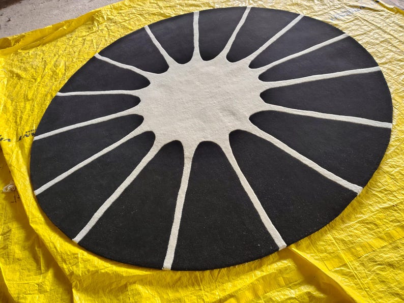 May include: A circular rug featuring a black background with a central white sunburst or star-like pattern. The white design consists of a solid circular center with long, tapered rays extending outward toward the edge of the rug.
