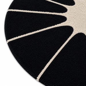 May include: A circular rug featuring a high-pile, tufted texture. The design consists of a solid black background with a cream-colored sunburst pattern radiating from the center, creating distinct, elongated, petal-like shapes.