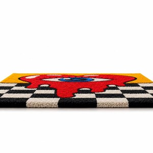May include: A rectangular tufted rug featuring a central red melting heart graphic with a large blue eye in the middle. The background consists of a yellow upper section and a black and white checkered pattern along the bottom edge of the rug.