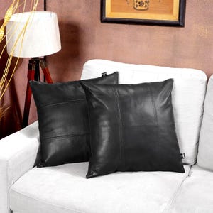 Könnte beinhalten: Zwei schwarze Lederkissen mit Steppdetails liegen auf einem weißen Sofa. Die Kissen haben eine quadratische Form und eine glatte, matte Oberfläche. Die Kissen sind einfarbig schwarz.