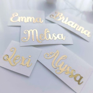 Op de afbeelding: Vijf witte rechthoekige naamlabels met gouden cursieve letters. De namen "Emma", "Brianna", "Melissa", "Lexi" en "Alyssa" worden weergegeven. De labels zijn gerangschikt op een wit oppervlak.