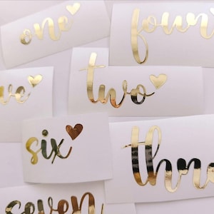 May include: Gold foil table number stickers on white rectangular labels. Numbers one to seven are written in a cursive font, each with a heart detail. Suitable for wedding or event decoration.