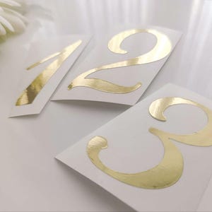May include: Gold metallic numbers 1, 2, and 3 on white cardstock. The numbers have a shiny, reflective surface and are arranged at different angles. The background is a soft white, with a hint of floral elements.