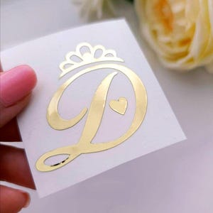May include: A gold-coloured sticker featuring the letter "D" with a heart and a crown design. The sticker is on a white background. The sticker is likely used for decoration or personalization.