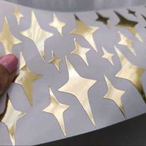 May include: A sheet of adhesive stickers featuring multiple four-pointed star shapes in a metallic gold finish. The stars vary in size and orientation, displaying a reflective surface against a plain white backing paper, arranged in a scattered pattern.