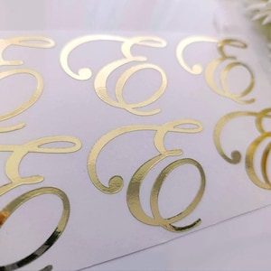 May include: A sheet of white adhesive backing featuring six individual gold foil stickers, each displaying the uppercase letter E in a decorative, flowing cursive script with elegant loops and a reflective, metallic finish.