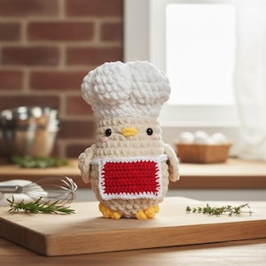 May include: A crocheted chef chicken doll wearing a white chef's hat and a red apron. The doll has a beige body, yellow feet, and small black eyes. It is standing on a wooden cutting board, with a brick wall in the background.