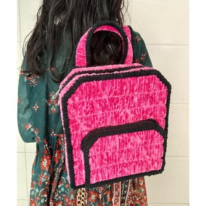 Könnte beinhalten: Ein leuchtend pinkfarbener Rucksack mit einem strukturierten, gerafften Design und schwarzem Besatz. Der Rucksack hat einen oberen Griff und eine Vordertasche. Die Tasche ist aus einem weichen, plüschigen Material gefertigt und wird auf dem Rücken getragen.