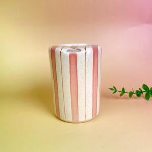 May include: A cylindrical ceramic vase with a cream-colored base and vertical pink stripes. The vase has a small opening at the top and is suitable for holding flowers or other decorative items. The design is simple and modern.