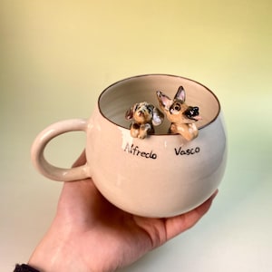 May include: Cream-colored ceramic mug with two dog figurines inside. One is a small, spotted dog, and the other is a German Shepherd. The names "Alfredo" and "Vasco" are written on the mug.