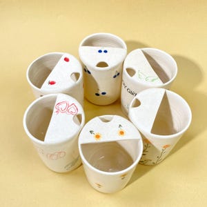 May include: A collection of six white ceramic cups with a textured finish. Each cup features a unique hand-painted design, including cherries, strawberries, and floral patterns. The cups have a distinctive half-moon cutout on the rim.