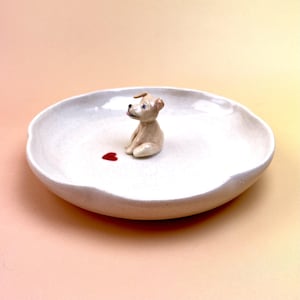 Peut inclure: Un plat en c&eacute;ramique blanc avec un bord ondul&eacute;, orn&eacute; d'une petite figurine de chiot beige et d'un c&oelig;ur rouge. Le chiot est assis et le c&oelig;ur est peint sur la surface du plat. Le fond est de couleur p&ecirc;che.