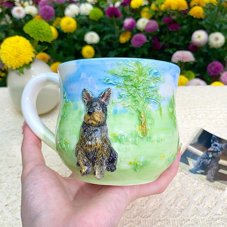 May include: A ceramic mug with a hand-painted landscape scene featuring a dog. The mug has a white handle and a detailed relief of a dog on a green and blue background. The mug is held in a hand.