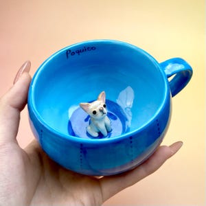 May include: A vibrant blue ceramic mug featuring a small, white chihuahua figurine inside. The mug has a handle and the word "Poquito" is written on the rim. The interior has a blue swirl design.