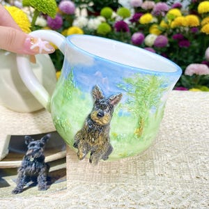 May include: A ceramic mug with a hand-painted landscape scene featuring a dog. The mug has a white handle and a blue rim. The dog is brown and black, and the background is green and blue.
