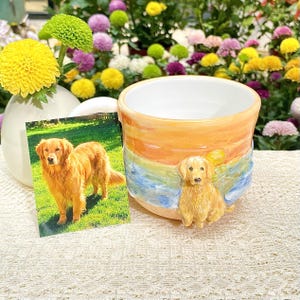 May include: A ceramic mug with a golden retriever design. The mug features a painted sunset scene with a sculpted dog. A photograph of a golden retriever is next to the mug. Flowers are in the background.
