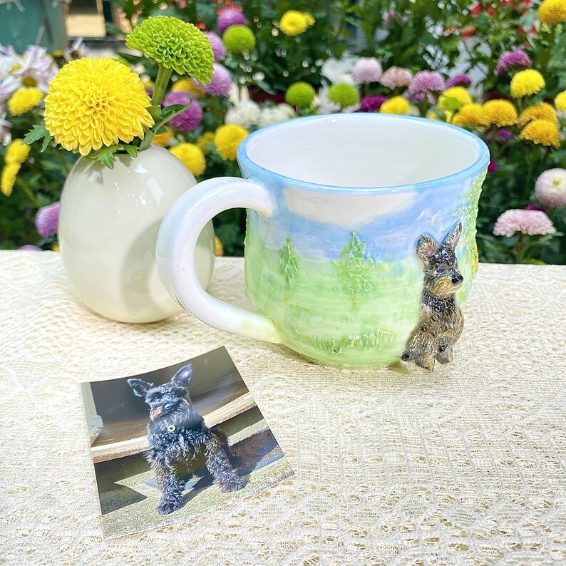 May include: A ceramic mug with a landscape design featuring a dog figurine. The mug has a white handle and a blue rim. A small vase with yellow flowers is next to the mug. A photo of a dog is in front of the mug.