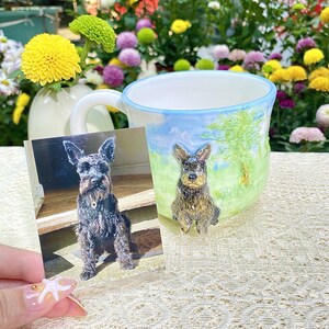 May include: A ceramic mug with a hand-painted landscape scene and a 3D dog figurine. A photograph of a dog is held next to the mug. The mug has a blue rim and a white handle. The background features colorful flowers.