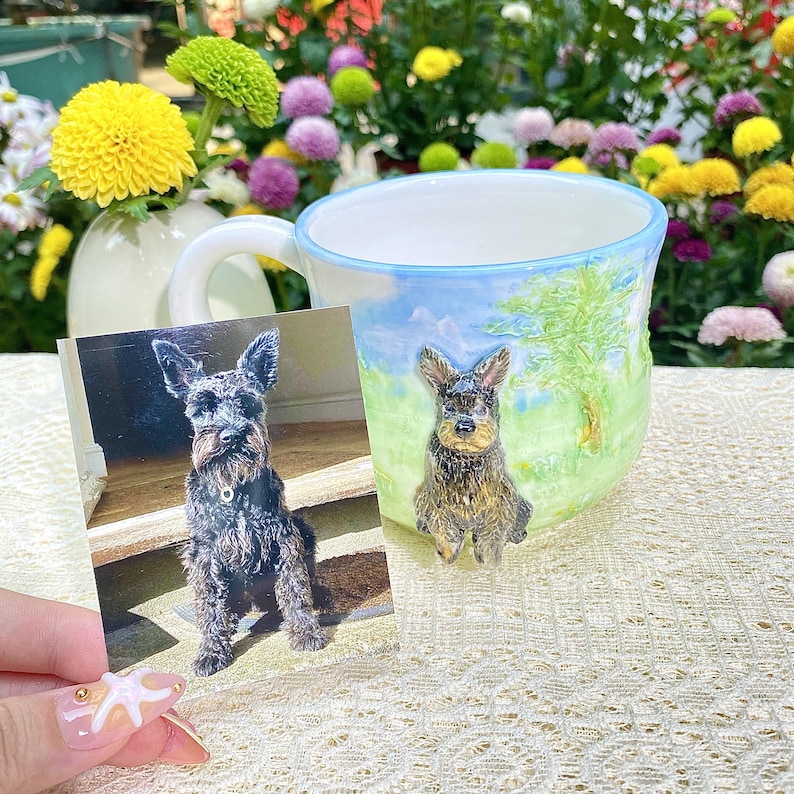 May include: A ceramic mug with a hand-painted landscape scene and a 3D dog figurine. A photograph of a dog is held next to the mug. The mug has a blue rim and a white handle. The background features colorful flowers.