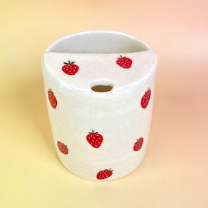 May include: A white ceramic wall-mounted container with a speckled finish, adorned with red strawberry illustrations. The container has a curved top with a hole and is designed for holding items. The overall design is simple and charming.