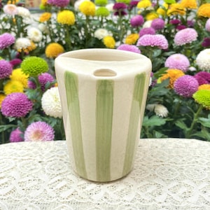 May include: A cream ceramic vase with vertical green stripes. The vase has a rounded shape and a small opening at the top. The background features a variety of colourful flowers.