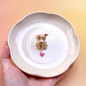 Peut inclure: Petite assiette en c&eacute;ramique blanc cass&eacute; &agrave; bord festonn&eacute;. Une figurine de chiot brun clair est au centre, au-dessus d'un c&oelig;ur rose. L'assiette est tenue dans une main sur un fond jaune et rose.
