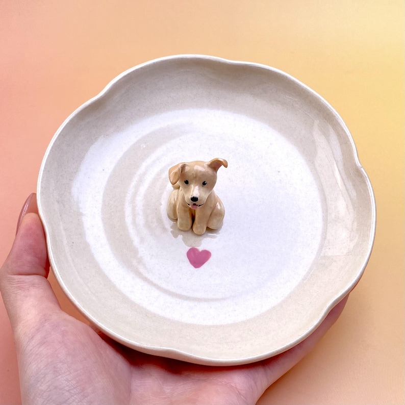Peut inclure: Petite assiette en c&eacute;ramique blanc cass&eacute; &agrave; bord festonn&eacute;. Une figurine de chiot brun clair est au centre, au-dessus d'un c&oelig;ur rose. L'assiette est tenue dans une main sur un fond jaune et rose.