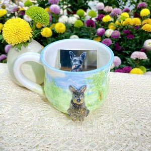 May include: A ceramic mug with a light blue rim and handle, featuring a hand-painted landscape design with a sculpted dog. The mug contains a small photo of a dog. The mug is set against a backdrop of colorful flowers.