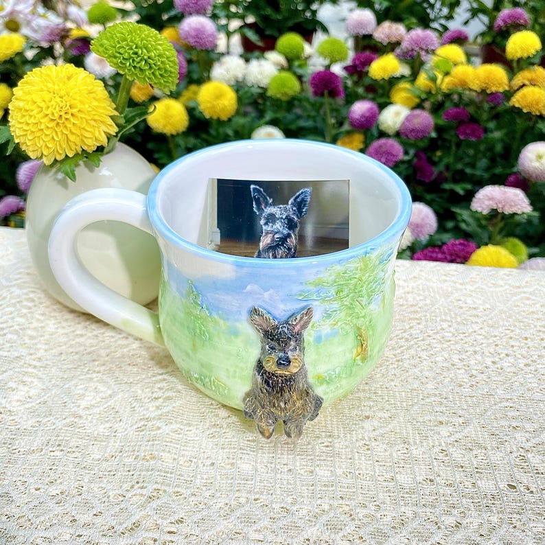 May include: A ceramic mug with a light blue rim and handle, featuring a hand-painted landscape design with a sculpted dog. The mug contains a small photo of a dog. The mug is set against a backdrop of colorful flowers.