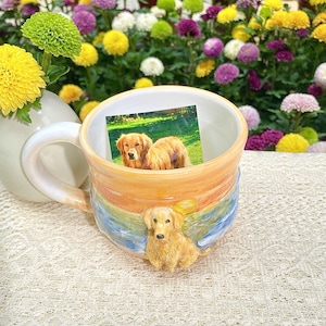 May include: A ceramic mug with a golden retriever theme. The mug features a sculpted golden retriever on the side and a photo of two golden retrievers inside. The mug has a colorful, layered design with orange, blue, and yellow hues.