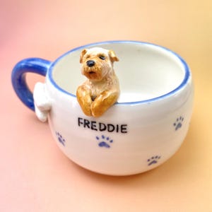 May include: A white ceramic mug with a blue handle and rim, featuring a 3D figurine of a tan dog. The mug is decorated with blue paw prints and the name "FREDDIE" in black lettering.