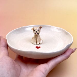 Peut inclure: Un petit plat en c&eacute;ramique blanc cass&eacute; avec un bord festonn&eacute;, orn&eacute; d'une figurine miniature de chien beige et d'un c&oelig;ur rouge. Le plat est tenu dans une main sur un fond d&eacute;grad&eacute; doux.