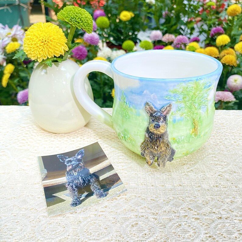 May include: A ceramic mug with a hand-painted landscape scene featuring a dog figurine. The mug has a light blue rim and a white handle. A small photo of the dog is next to the mug. A small white vase holds yellow and green flowers.