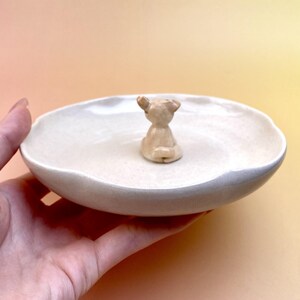 Peut inclure: Un petit plat en c&eacute;ramique blanc cass&eacute; avec un bord ondul&eacute;, orn&eacute; d'une minuscule figurine d'ours brun clair au centre. Le plat est tenu dans une main sur un fond jaune-orang&eacute; doux. L'ours est en position assise.