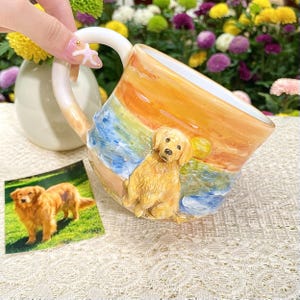 May include: A ceramic mug featuring a golden retriever on a beach scene with a sunset design. The mug has a white handle and a matching small photo of a golden retriever. The mug is held by a person.
