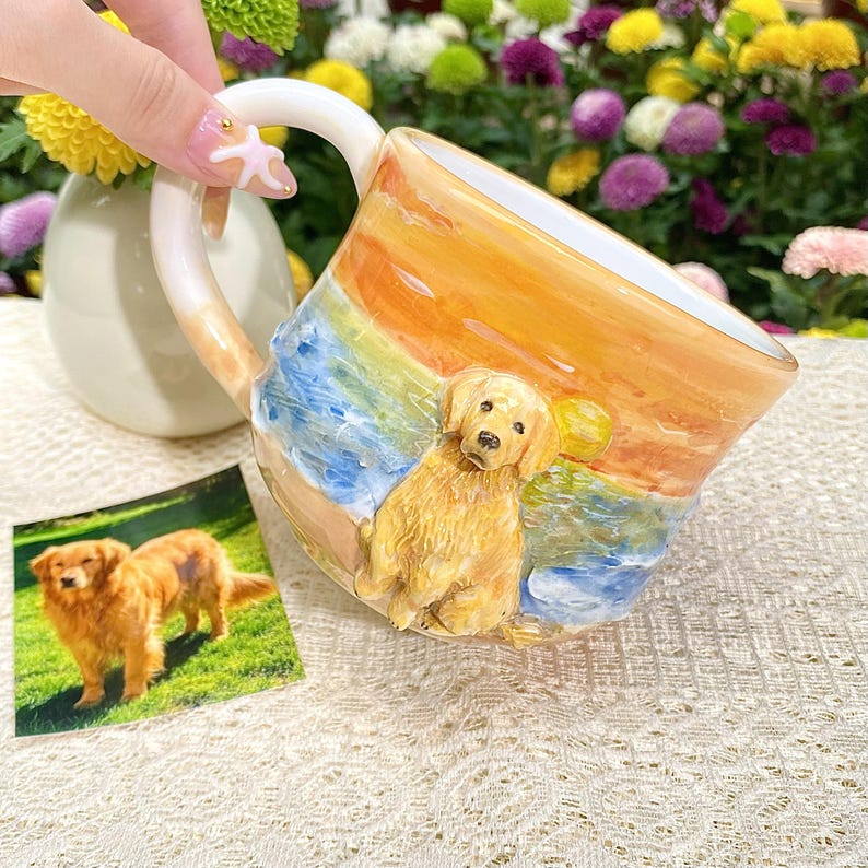 May include: A ceramic mug featuring a golden retriever on a beach scene with a sunset design. The mug has a white handle and a matching small photo of a golden retriever. The mug is held by a person.